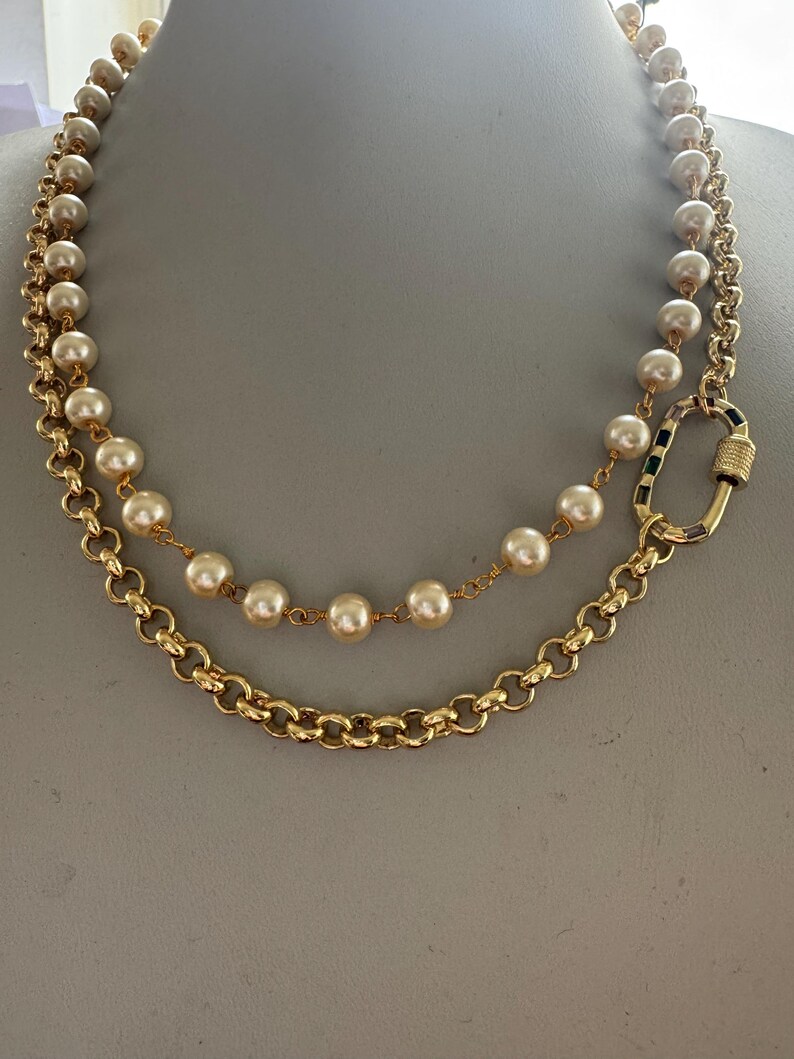 May include: A gold-toned necklace with two strands. The top strand features cream-colored pearls, while the bottom strand is a chunky chain. The necklace is fastened with a gold clasp with black and green accents.