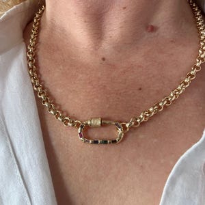 May include: Gold-toned chain necklace with a large, decorative clasp. The clasp is adorned with a row of colorful gemstones. The necklace is worn against a white shirt.