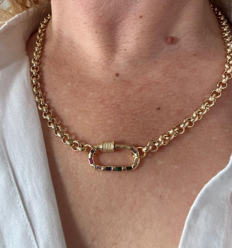 May include: Gold-toned chain necklace with a large, decorative clasp. The clasp is adorned with a row of colorful gemstones. The necklace is worn against a white shirt.