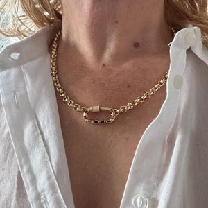 May include: A gold-colored chain necklace with a large, oval-shaped clasp. The clasp is adorned with small, colorful gemstones. The necklace is worn over a white button-down shirt.