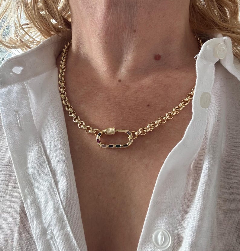 May include: A gold-colored chain necklace with a large, oval-shaped clasp. The clasp is adorned with small, colorful gemstones. The necklace is worn over a white button-down shirt.