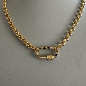 May include: A gold-colored chain necklace with a large, decorative clasp. The chain links are a rounded, curb style. The clasp is a rectangular shape with colorful, small gemstones. The necklace is displayed on a neutral background.