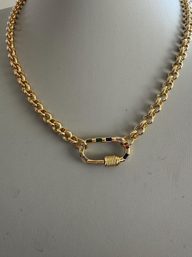 May include: A gold-colored chain necklace with a large, decorative clasp. The chain links are a rounded, curb style. The clasp is a rectangular shape with colorful, small gemstones. The necklace is displayed on a neutral background.