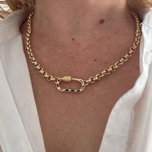 May include: Gold-toned necklace with a large, colorful clasp. The chain is made of rounded links. The clasp features a rectangular design with a variety of colored stones. The necklace is worn against a white shirt.