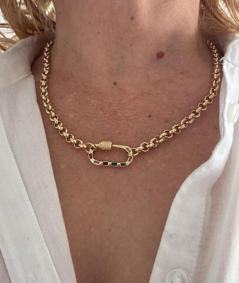 May include: Gold-toned necklace with a large, colorful clasp. The chain is made of rounded links. The clasp features a rectangular design with a variety of colored stones. The necklace is worn against a white shirt.