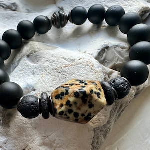 Peut inclure: Bracelet en perles noires avec une grosse perle de pierre géométrique à motifs marrons et blancs. Le bracelet comporte également de petites perles de lave noire et des accents métalliques argentés.