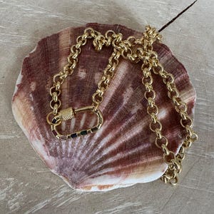 May include: A gold-colored necklace with a large carabiner-style clasp featuring a row of colorful stones. The necklace is composed of a chain of rounded links. The jewelry rests on a large, textured seashell with brown and tan hues.