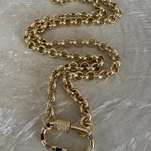 May include: Gold-toned necklace with a large carabiner clasp. The clasp is adorned with colorful gemstones. The chain is made of interlocking oval links. The necklace is displayed on a light gray surface.
