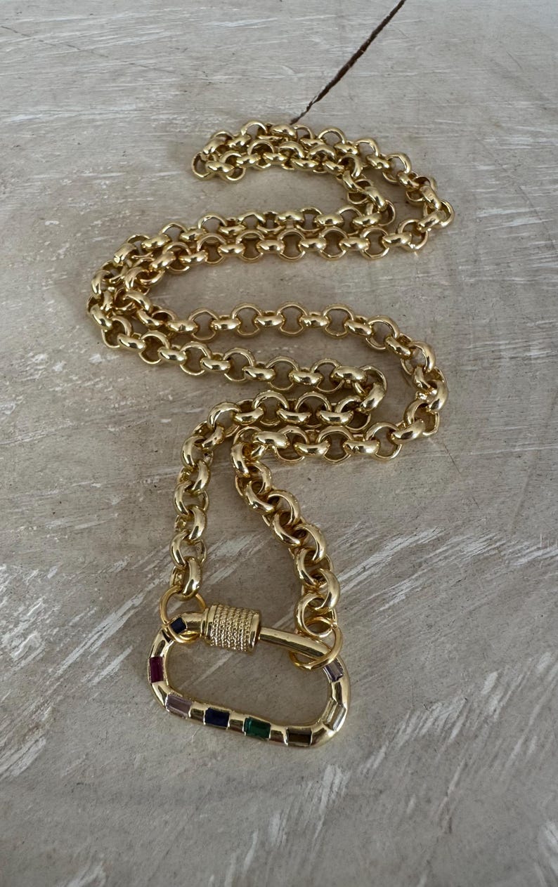 May include: Gold-toned necklace with a large carabiner clasp. The clasp is adorned with colorful gemstones. The chain is made of interlocking oval links. The necklace is displayed on a light gray surface.