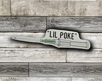 May include: A sticker featuring a syringe design with the text "LIL POKE" in black. The syringe has a green tip and a clear body with black detailing. The sticker is on a wooden background.