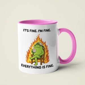 May include: White ceramic mug with a pink interior and handle. The mug features a cartoon frog sitting in a chair surrounded by flames, holding a coffee cup. Text on the mug reads "IT'S FINE. I'M FINE. EVERYTHING IS FINE."