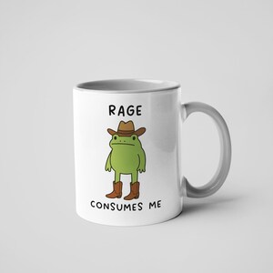 May include: White ceramic mug with a grey handle. It features a cartoon frog in a cowboy hat and boots, with the text "RAGE CONSUMES ME". Suitable for hot or cold drinks, this mug is a fun, quirky item.