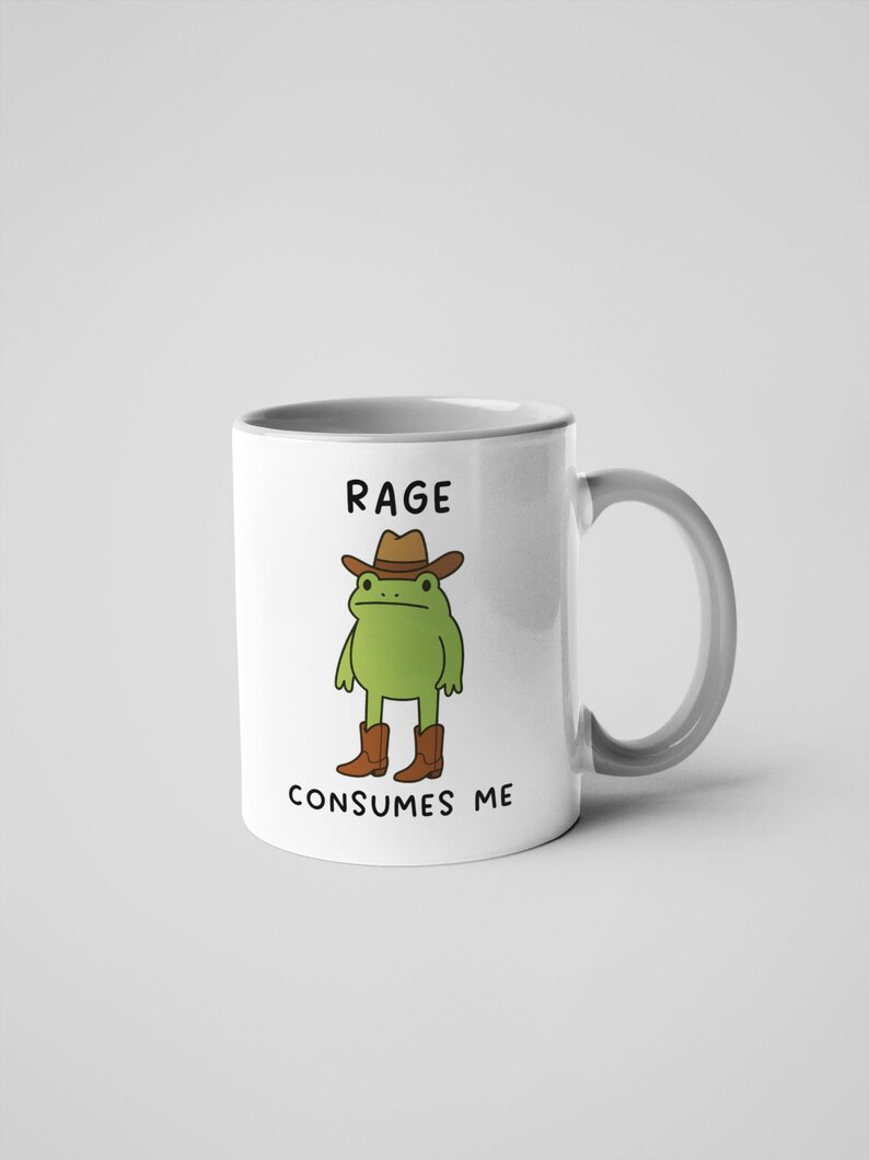 May include: White ceramic mug with a grey handle. It features a cartoon frog in a cowboy hat and boots, with the text "RAGE CONSUMES ME". Suitable for hot or cold drinks, this mug is a fun, quirky item.