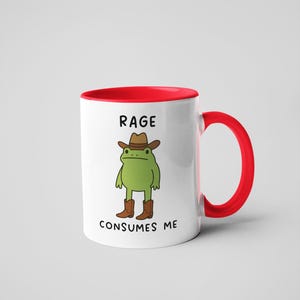 May include: White ceramic mug with a red interior and handle. The mug features a cartoon frog wearing a cowboy hat and boots, with the text "RAGE CONSUMES ME" in black.