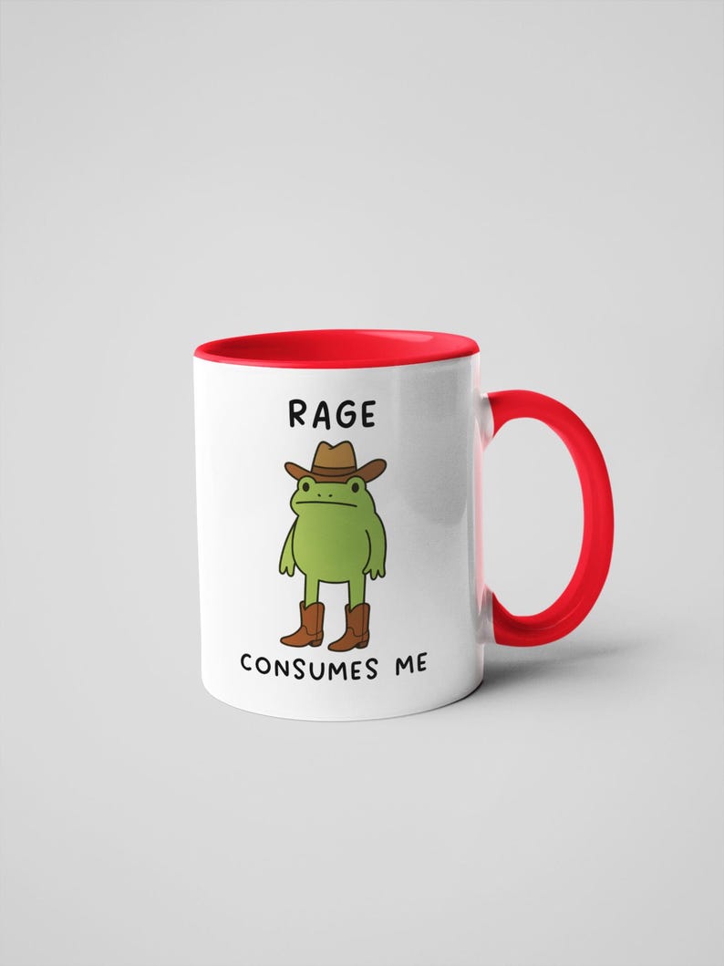 May include: White ceramic mug with a red interior and handle. The mug features a cartoon frog wearing a cowboy hat and boots, with the text "RAGE CONSUMES ME" in black.
