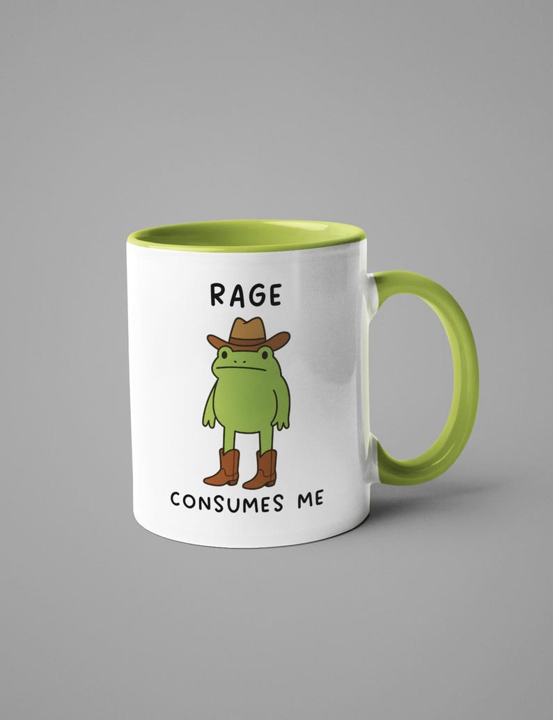 May include: White ceramic mug with a green interior and handle. The mug features a cartoon frog wearing a cowboy hat and boots. Text on the mug reads "RAGE CONSUMES ME."