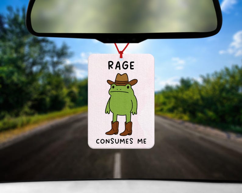 May include: A rectangular car air freshener with a cartoon frog wearing a cowboy hat and boots. The text "RAGE CONSUMES ME" is printed above and below the frog illustration. The air freshener hangs from a red string.