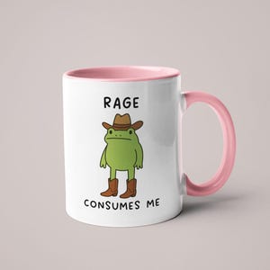 May include: White ceramic mug with a pink interior and handle. The mug features a cartoon frog wearing a cowboy hat and boots. The text "RAGE CONSUMES ME" is printed on the mug.