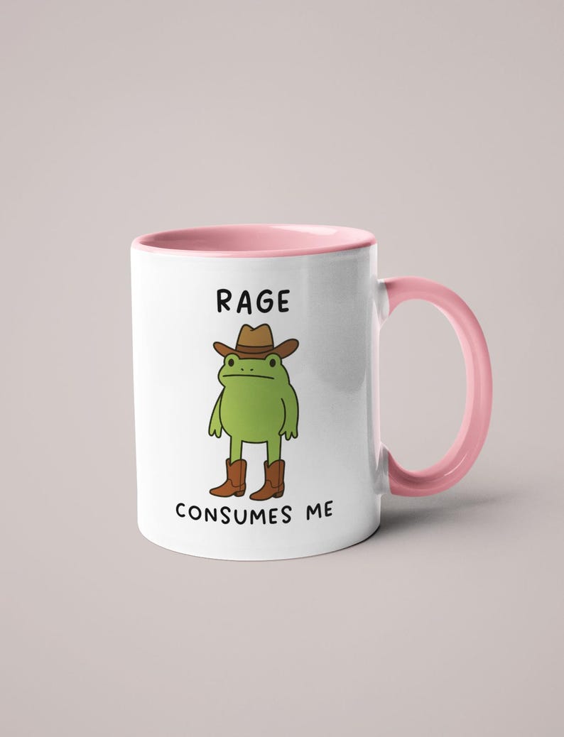 May include: White ceramic mug with a pink interior and handle. The mug features a cartoon frog wearing a cowboy hat and boots. The text "RAGE CONSUMES ME" is printed on the mug.
