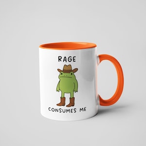 May include: White ceramic mug with an orange interior and handle. The mug features a cartoon frog wearing a cowboy hat and boots. The text "RAGE CONSUMES ME" is printed on the mug.