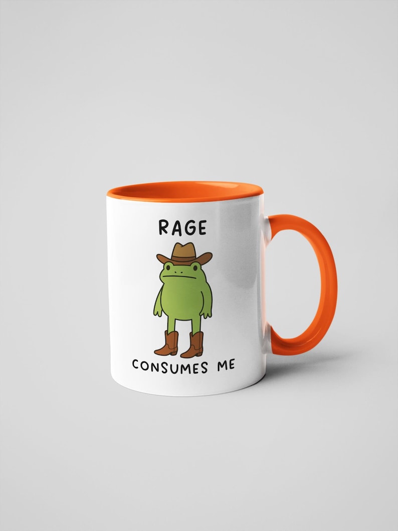 May include: White ceramic mug with an orange interior and handle. The mug features a cartoon frog wearing a cowboy hat and boots. The text "RAGE CONSUMES ME" is printed on the mug.