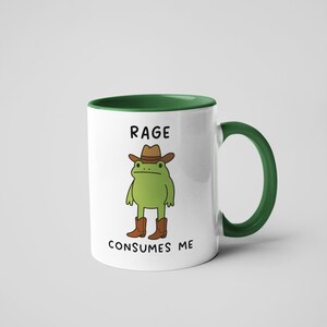 May include: White ceramic mug with a green interior and handle. The mug features a cartoon frog wearing a cowboy hat and boots. The text "RAGE CONSUMES ME" is printed on the mug.