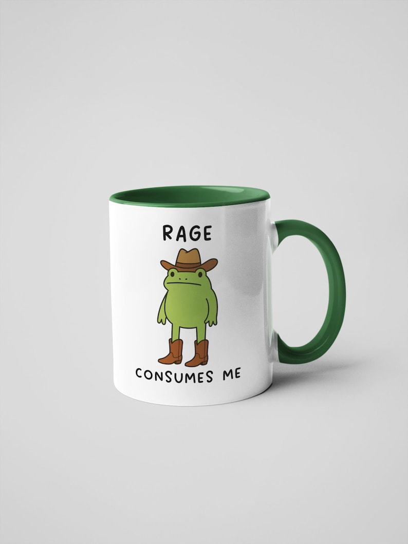 May include: White ceramic mug with a green interior and handle. The mug features a cartoon frog wearing a cowboy hat and boots. The text "RAGE CONSUMES ME" is printed on the mug.