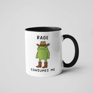 May include: White ceramic mug with a black interior and handle. The mug features a cartoon frog wearing a cowboy hat and boots. The text on the mug reads "RAGE CONSUMES ME".