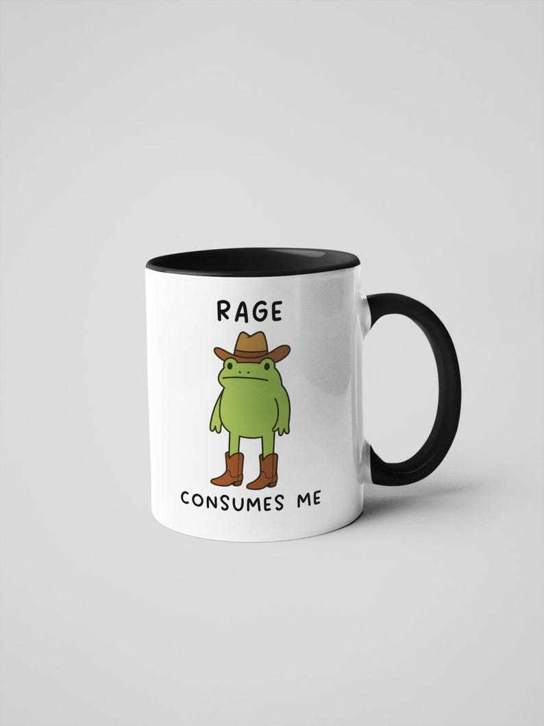 May include: White ceramic mug with a black interior and handle. The mug features a cartoon frog wearing a cowboy hat and boots. The text on the mug reads "RAGE CONSUMES ME".
