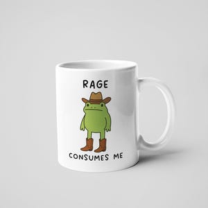 May include: White ceramic mug with a cartoon frog in a cowboy hat and boots. The mug has the text "RAGE CONSUMES ME". The frog is green, the hat and boots are brown. The mug is a standard size.