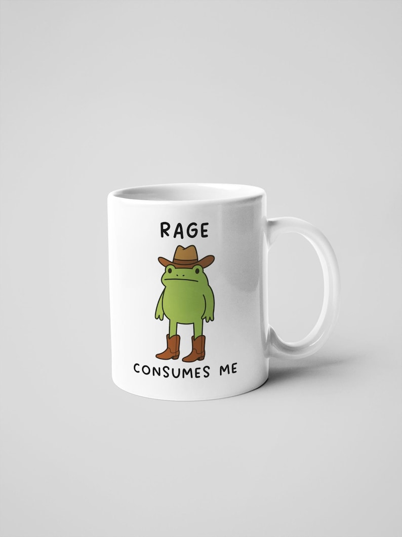 May include: White ceramic mug with a cartoon frog in a cowboy hat and boots. The mug has the text "RAGE CONSUMES ME". The frog is green, the hat and boots are brown. The mug is a standard size.