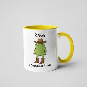 May include: White ceramic mug with a yellow interior and handle. The mug features a cartoon frog wearing a cowboy hat and boots. Text on the mug reads "RAGE CONSUMES ME."
