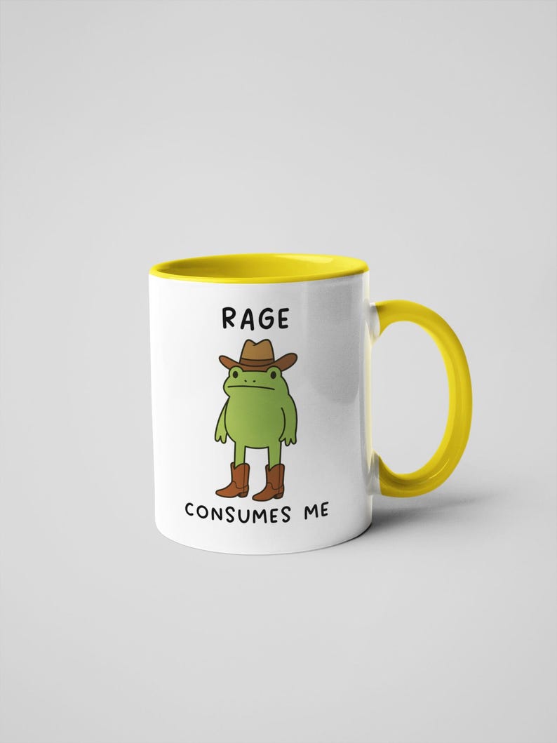 May include: White ceramic mug with a yellow interior and handle. The mug features a cartoon frog wearing a cowboy hat and boots. Text on the mug reads "RAGE CONSUMES ME."