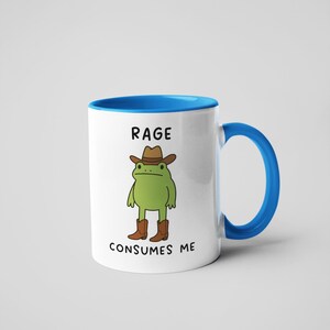 May include: White ceramic mug with a blue interior and handle. The mug features a cartoon frog wearing a cowboy hat and boots. The text "RAGE CONSUMES ME" is printed on the mug.
