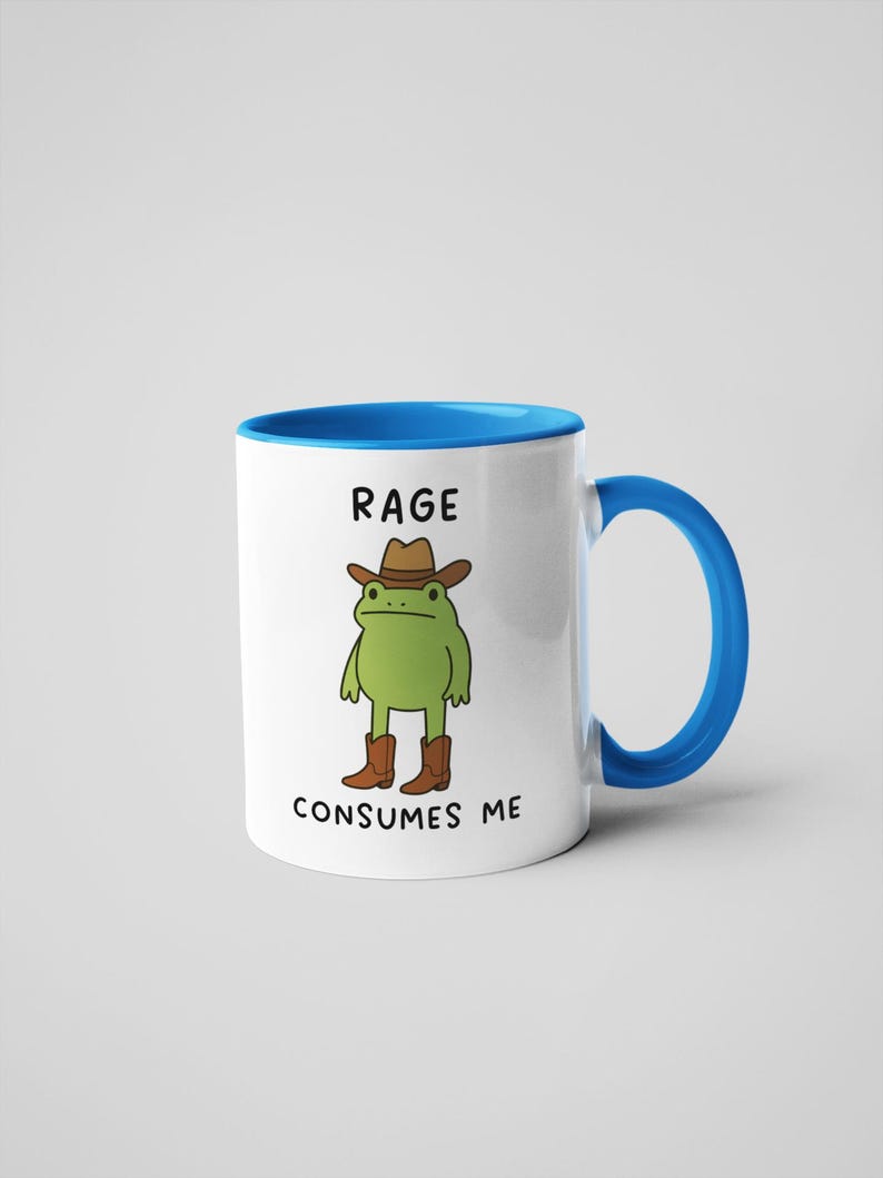 May include: White ceramic mug with a blue interior and handle. The mug features a cartoon frog wearing a cowboy hat and boots. The text "RAGE CONSUMES ME" is printed on the mug.