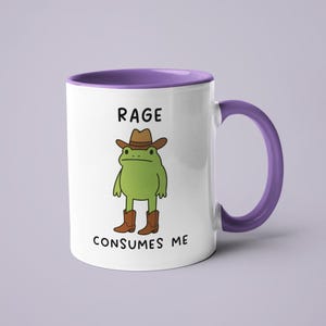 May include: White ceramic mug with a purple interior and handle. The mug features a cartoon frog wearing a cowboy hat and boots. Text on the mug reads "RAGE CONSUMES ME."