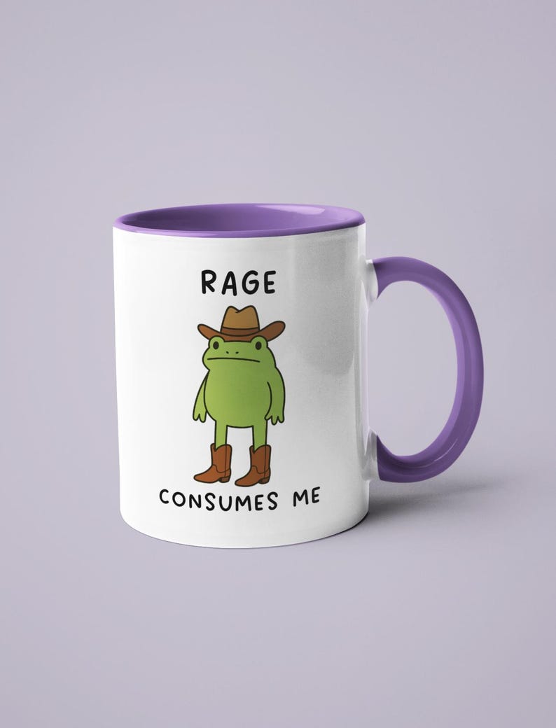 May include: White ceramic mug with a purple interior and handle. The mug features a cartoon frog wearing a cowboy hat and boots. Text on the mug reads "RAGE CONSUMES ME."
