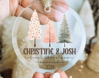 Könnte beinhalten: Transparenter Weihnachtsbaumschmuck aus Glas mit goldenem Band. Der Schmuck zeigt drei illustrierte Weihnachtsbäume in Rosa- und Grautönen. Der Text auf dem Schmuck lautet "CHRISTINE & JOSH first Christmas married 2024."
