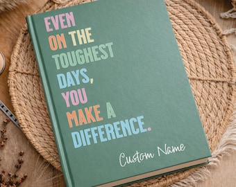 May include: A green journal with the text "EVEN ON THE TOUGHEST DAYS, YOU MAKE A DIFFERENCE." in colorful letters. The words "Custom Name" are written in cursive at the bottom. The journal is on a woven mat.