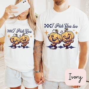 May include: A couple wearing matching white t-shirts with a vintage-style graphic of two dancing pumpkins. The shirts say "I Pick You" and "I Pick You Too" with checkered flags and musical notes. The shirts are ivory.