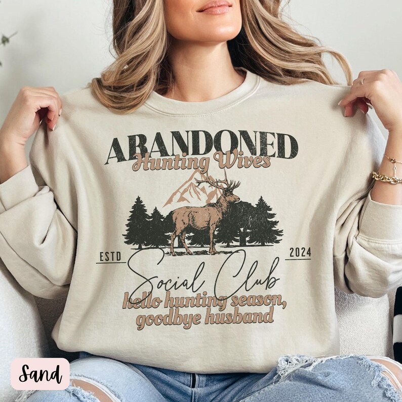 May include: A beige sweatshirt with the text "Abandoned Hunting Wives Social Club" and a deer illustration. The text also includes "Est. 2024" and "Hello hunting season, goodbye husband."