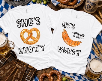 May include: Two white t-shirts featuring graphic prints. The left shirt displays a pretzel illustration with the text "SHE'S KNOTTY". The right shirt displays a sausage illustration with the text "HE'S THE WURST". Both shirts have short sleeves and crew necklines.
