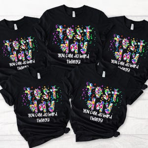Peut inclure: T-shirts noirs avec les mots "TEST DAY" en lettres colorées et pointillées. En dessous, la phrase "you can do hard things!" en blanc. Les t-shirts sont des vêtements décontractés.