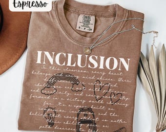 May include: Espresso-colored t-shirt with the word "INCLUSION" in large white letters. The shirt features a detailed message about inclusivity, with illustrations of a rainbow, heart, and backpack. A silver necklace with a pendant rests on the shirt.