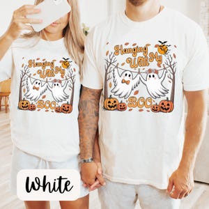 May include: White t-shirts with a Halloween-themed graphic. The design features two cartoon ghosts hanging from a clothesline, with pumpkins and the text "Hanging With My BOO". A black bat and orange sun are also included.