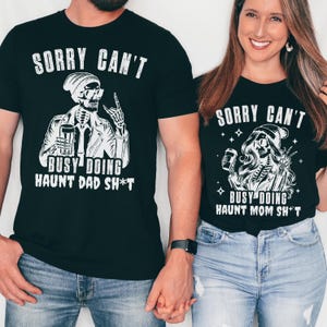 May include: Black t-shirts with white graphic designs. One shirt reads "SORRY CAN'T BUSY DOING HAUNT DAD SH*T" with a skeleton illustration. The other shirt says "SORRY CAN'T BUSY DOING HAUNT MOM SH*T" with a similar design. Both shirts are worn by a couple.