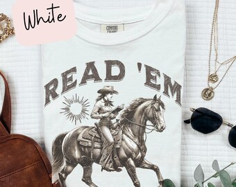 Peut inclure: T-shirt blanc avec un motif brun d'une femme à cheval lisant un livre. Le texte "READ 'EM" est imprimé en brun au-dessus du motif.