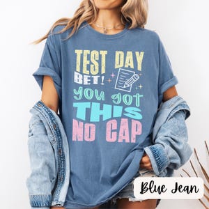 May include: A blue jean t-shirt with the text "Test Day Bet! You got this No Cap" in colorful letters. The text is stylized with a checkmark and pencil graphic.