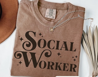 May include: Espresso-colored t-shirt with the words "Social Worker" in a decorative font. The shirt has a crew neck and short sleeves. A silver necklace with a pendant is draped across the shirt. A brown hat and dried plant are in the background.