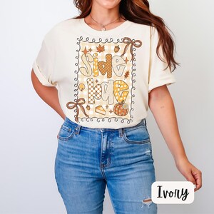 May include: Ivory-colored t-shirt with the words "Give Love" in a fall-themed design. The design includes a pumpkin, pie, and leaves. The shirt has short sleeves and a crew neck.
